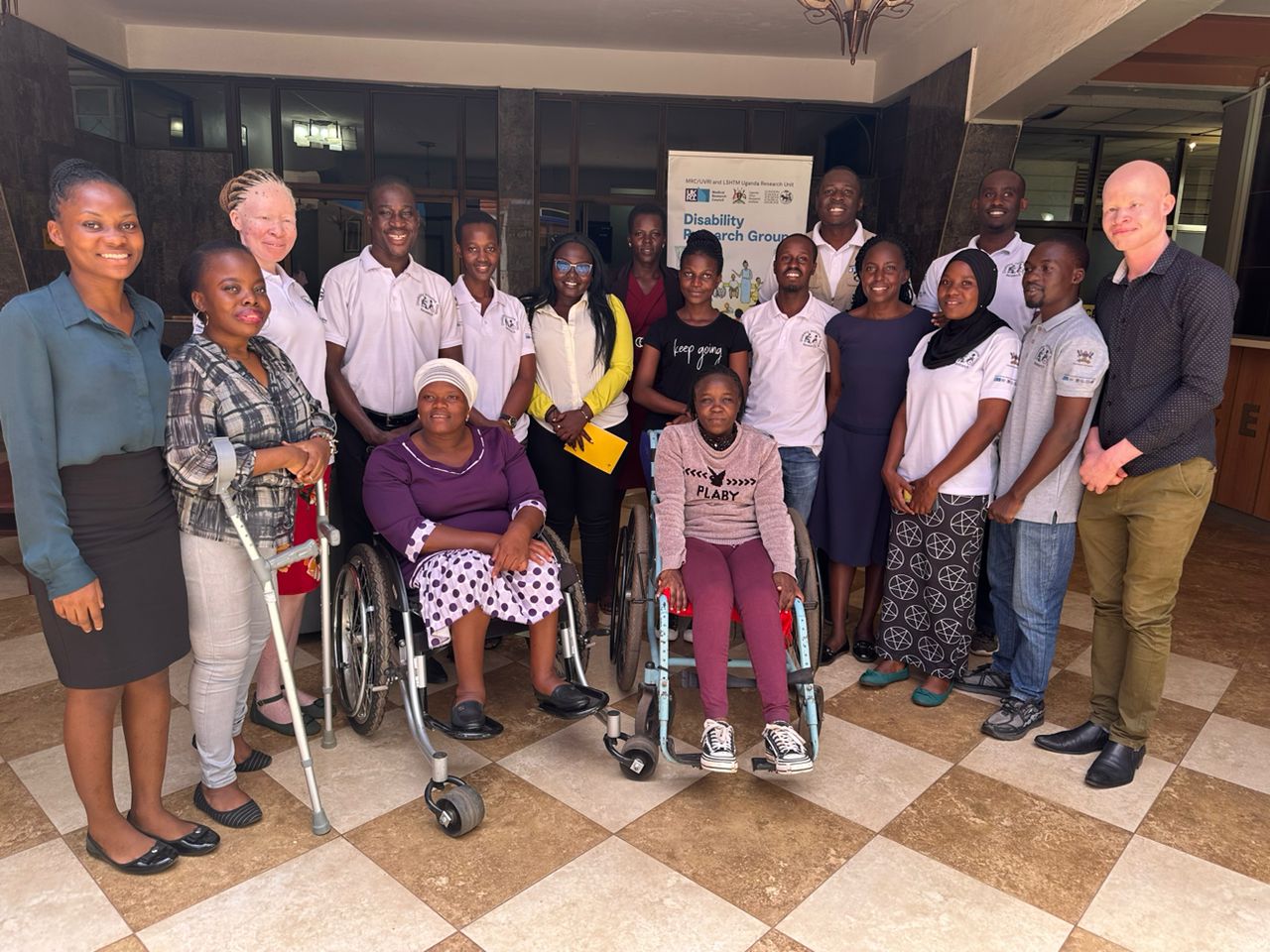 The image depicts a diverse group of individuals gathered together, likely for a meeting or event related to disability advocacy or research. The group includes both men and women, some of whom are using wheelchairs or crutches, highlighting a focus on inclusivity and representation of people with disabilities.   They are dressed in a mix of formal and casual attire, with some wearing matching shirts that suggest they are part of an organization or initiative. The setting appears to be indoors, with a banner in the background indicating a focus on disability research. The overall atmosphere seems positive and collaborative, reflecting a commitment to addressing issues related to disability.