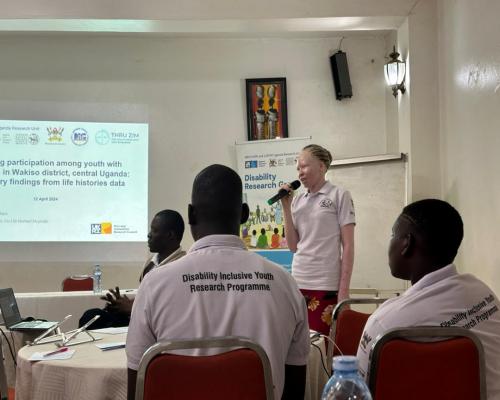 The image depicts a presentation taking place in a conference or workshop setting. A woman is speaking into a microphone, likely presenting findings related to youth participation among individuals with disabilities in Wakiso district, central Uganda. The presentation is supported by a slide displayed on a screen, which includes the title of the research and the date, April 12, 2024.  In the foreground, several attendees are seated at a table, wearing matching shirts that indicate their involvement in a "Di